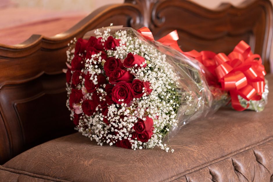 A bouquet arrangement of fresh red roses with velvety petals and green stems, accented by clusters of delicate white baby's breath flowers. The bouquet is wrapped in clear plastic and secured with a l