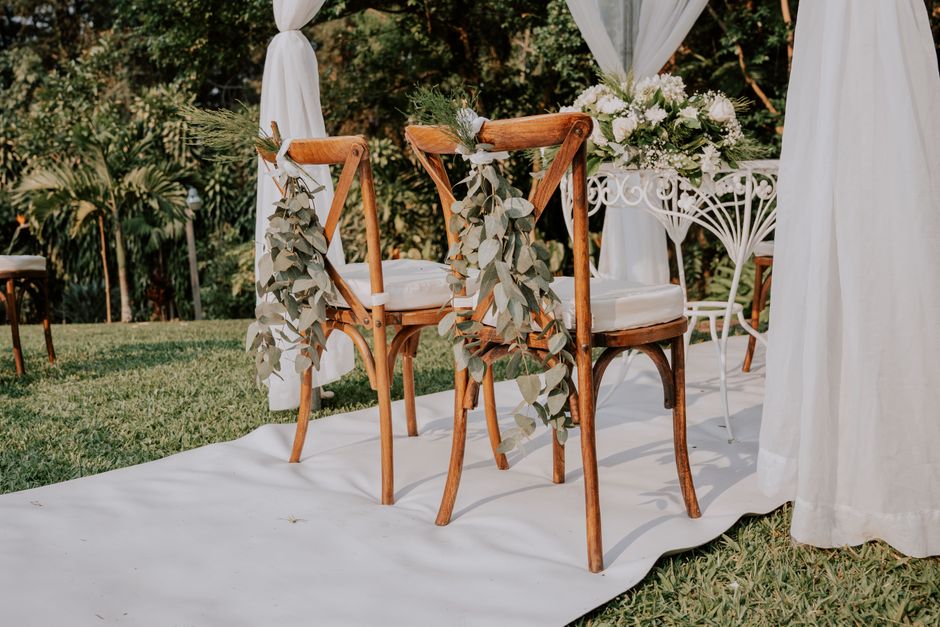 A floral arrangement featuring white roses and greenery placed on a white metal table with ornate detailing, set in an outdoor wedding venue. Two wooden chairs with white cushions are decorated with c