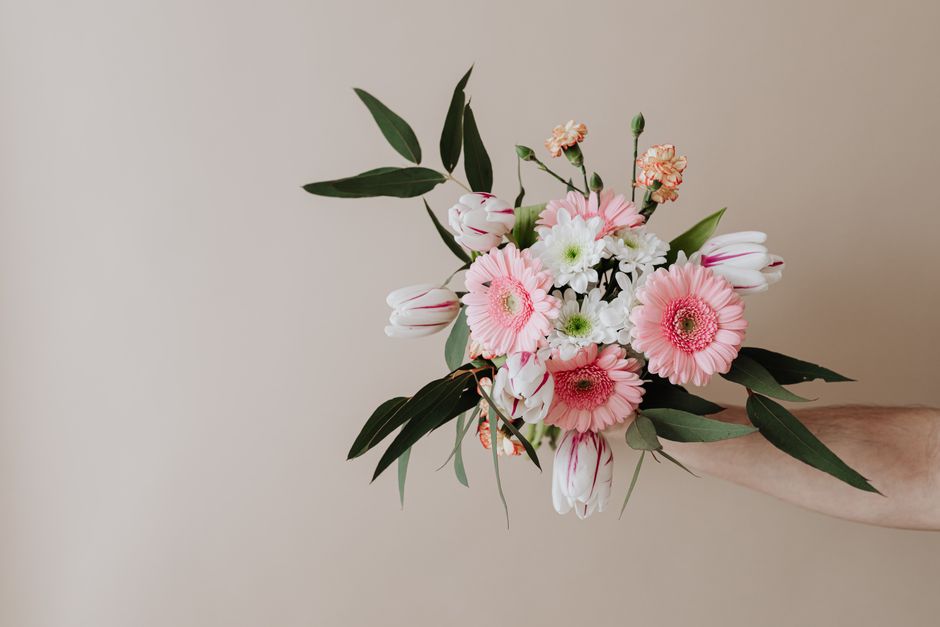 A hand holding a floral bouquet arranged with pink gerbera daisies, white daisies, tulips, and small pink carnations, complemented by green leafy foliage. The flowers are fresh with vibrant, soft peta