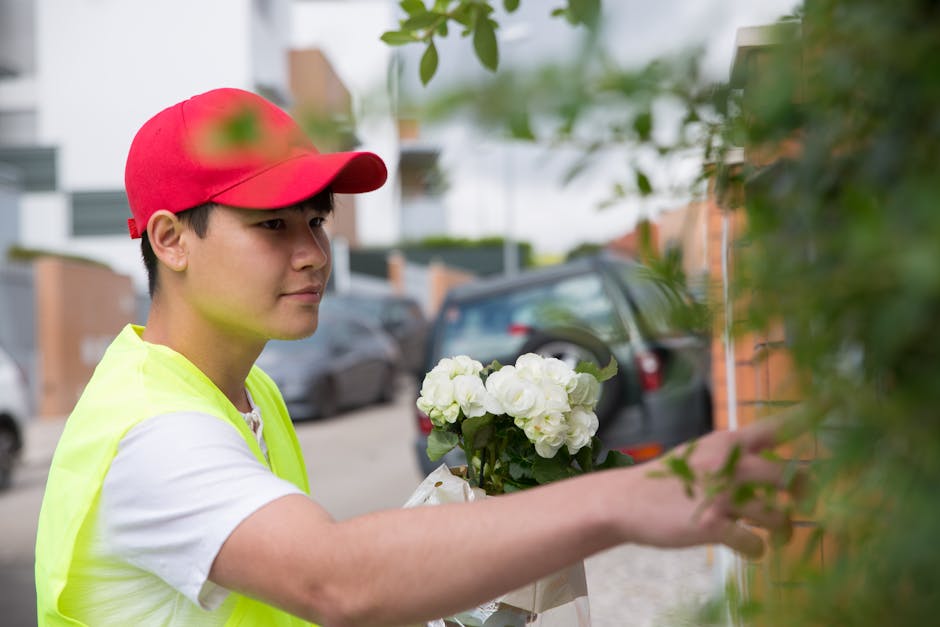 A young florist wearing a red cap and a bright yellow safety vest is arranging a bouquet of fresh white flowers, possibly hydrangeas or roses, in an outdoor setting near a brick wall. The flowers are