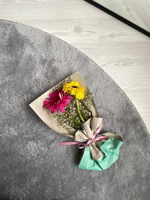 A small floral bouquet featuring vibrant pink and yellow daisies accented with baby's breath, wrapped in cream and light teal wrapping paper tied with a pink ribbon, resting on a grey textured surface